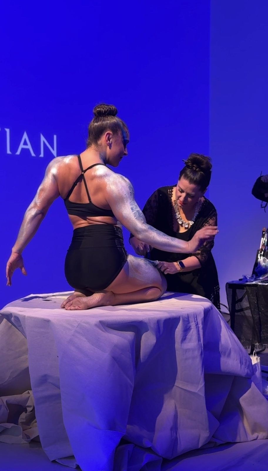 Body painter adding silver detail to a performer kneeling on a draped plinth under blue light
